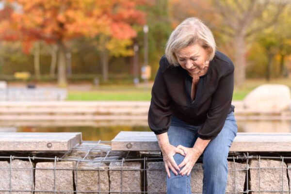 Elderly lady grabbing her knee in pain as she sits on a wall surrounding a pond after injuring herself out walking on an autumn day.