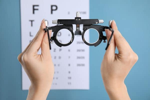 Close up image of a woman holding trial frame against eye chart test on blue background. 